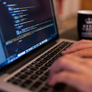 A close-up of a person's hands typing on a laptop keyboard with lines of code displayed on the screen. A black coffee mug with a white design is placed in the background.